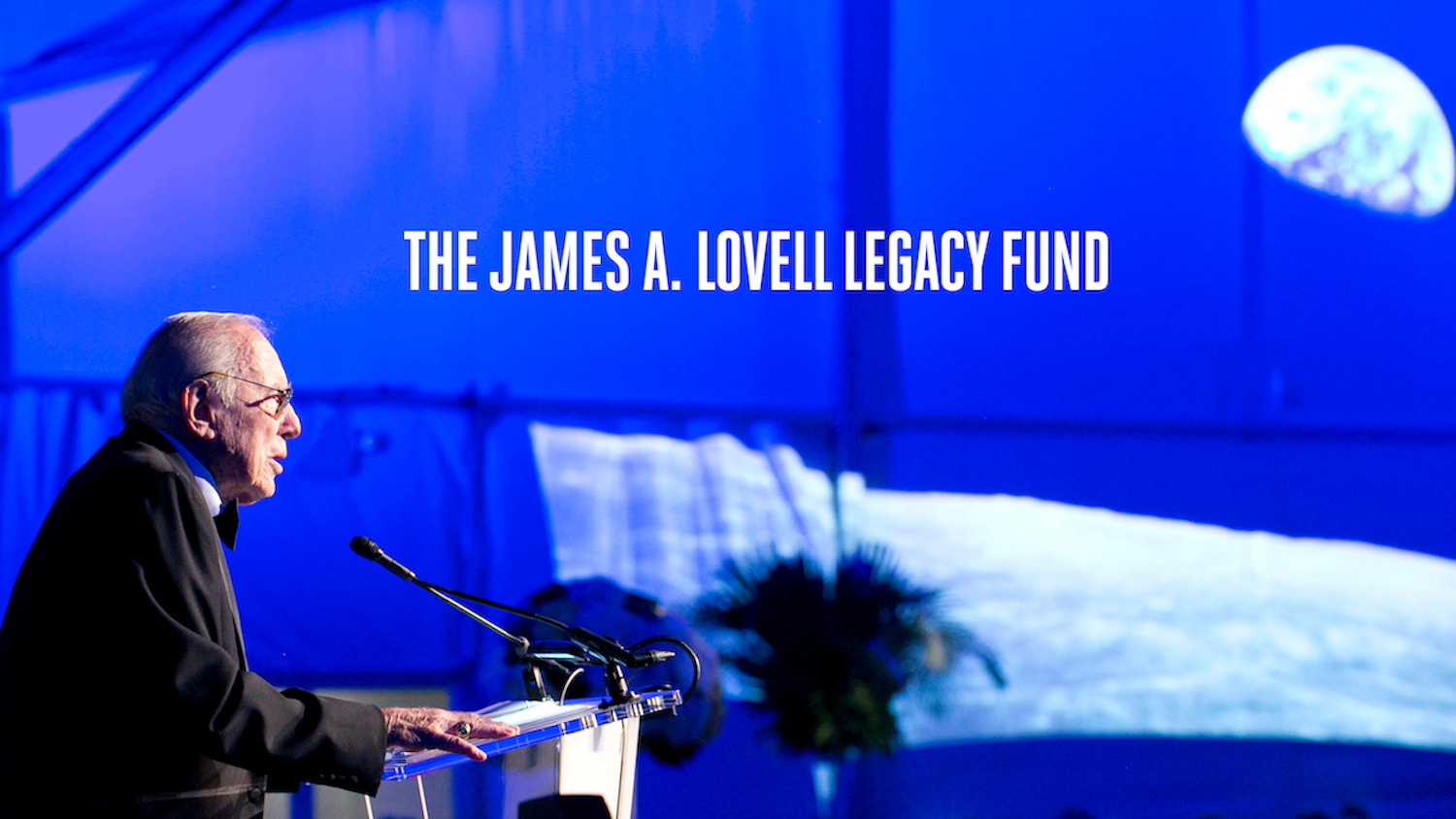 Capt Jim Lovell dressed in a tux stands at a podium accepting a Lifetime Achievement Award at the Adler. The Words "The James A. Lovell Legacy Fund" sit against a backdrop of the famous "Earthrise" photo.