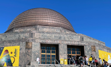 The Adler Planetarium on a sunny, summer day, with a line of guests on the museum steps. A bright blue sky contrasts the museum's yellow banners.
