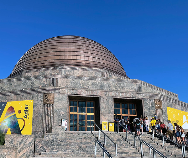 The Adler Planetarium on a sunny, summer day, with a line of guests on the museum steps. A bright blue sky contrasts the museum's yellow banners.