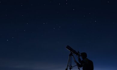 A silhouette of a person kneeling in front of a telescope, aimed at the night sky. The background shows a dark blue night sky with dim stars in the distance.