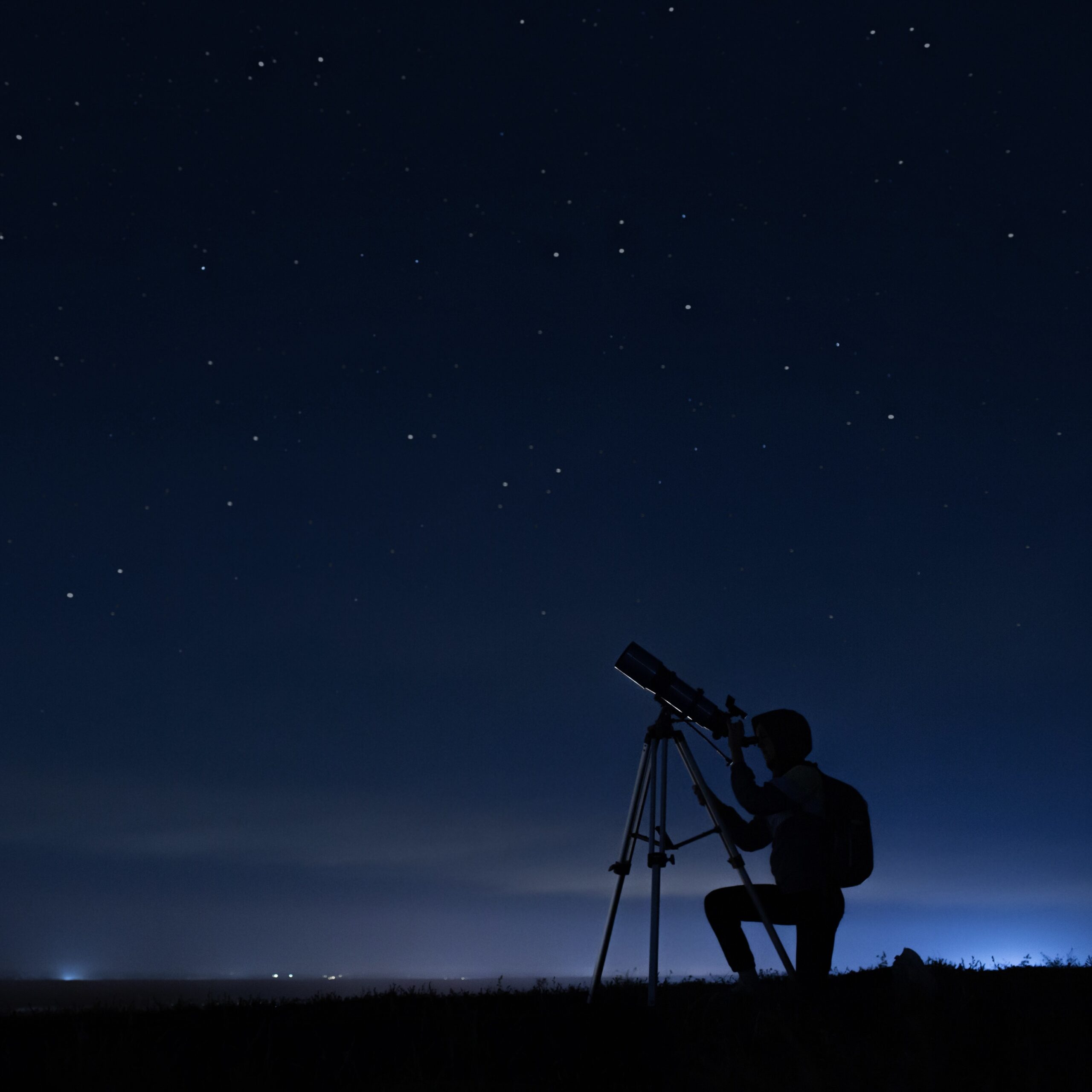 A silhouette of a person kneeling in front of a telescope, aimed at the night sky. The background shows a dark blue night sky with dim stars in the distance.